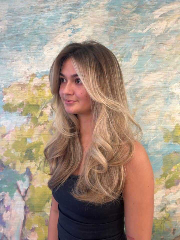 Woman with long, wavy blonde hair stands against a colorful floral background.