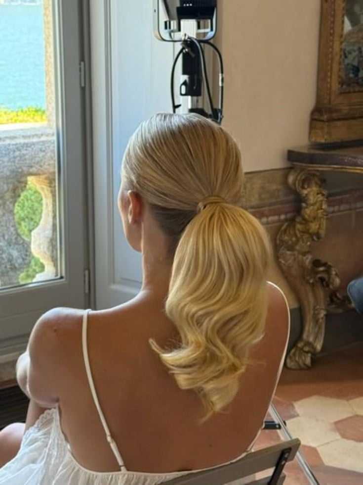 Blonde woman with wavy ponytail, sitting indoors near a window with lake view.