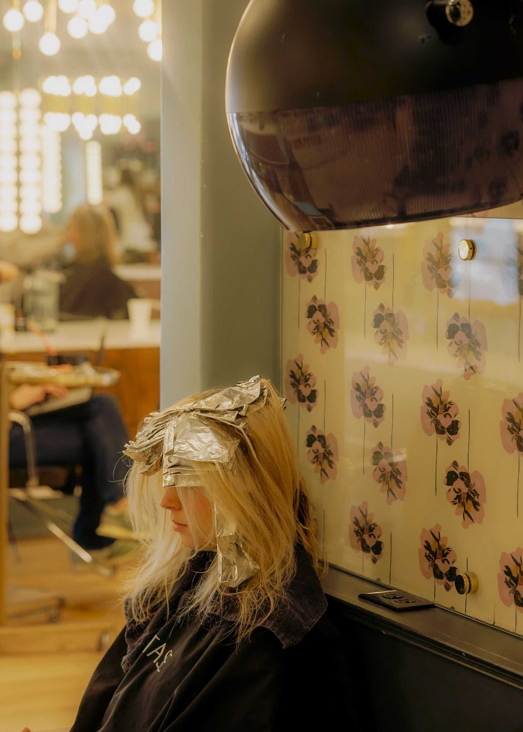 Person with foils in hair sits under salon dryer, surrounded by mirrors and lights.
