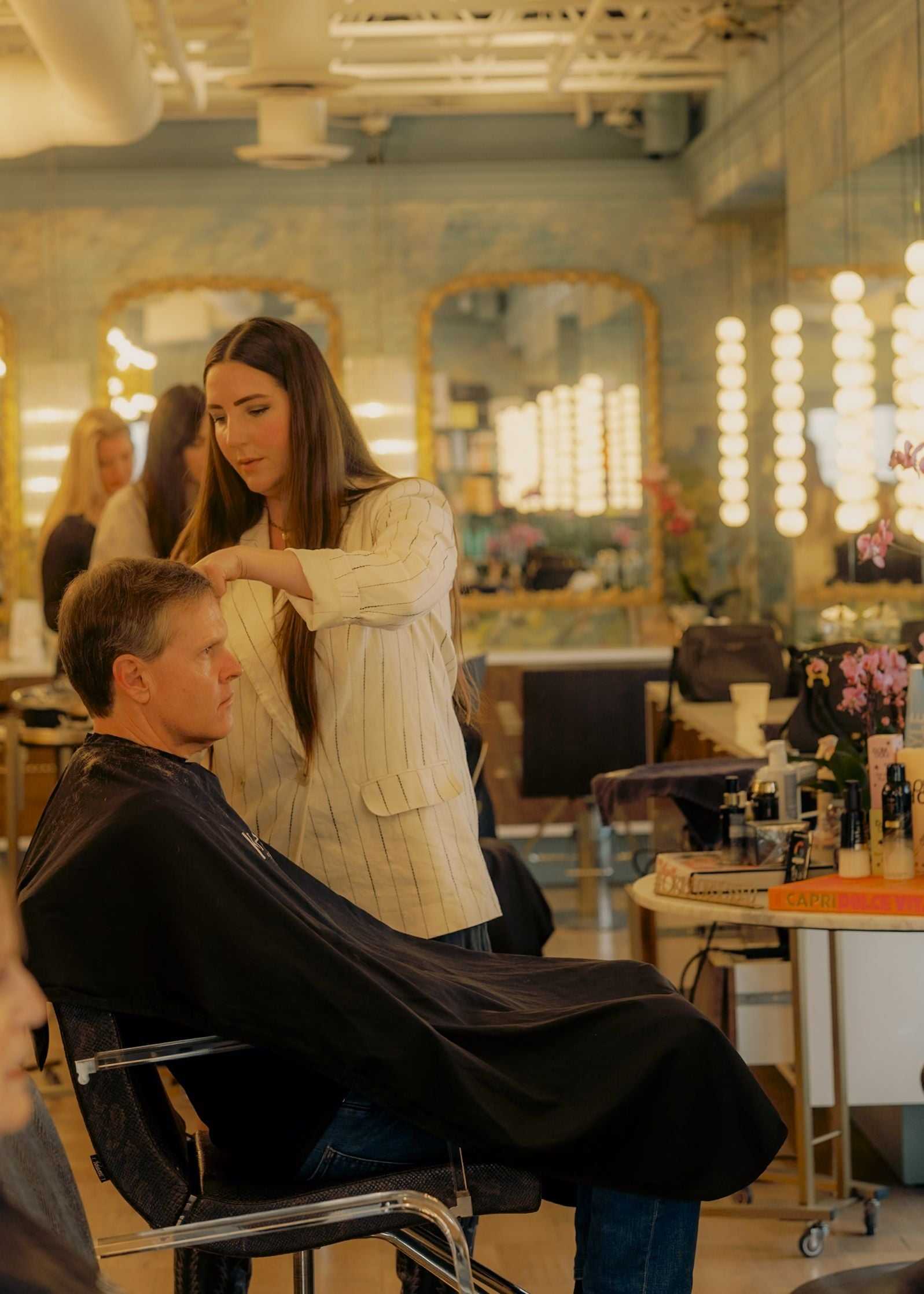 A stylist trims a man's hair in a salon with mirrors and bright lighting.