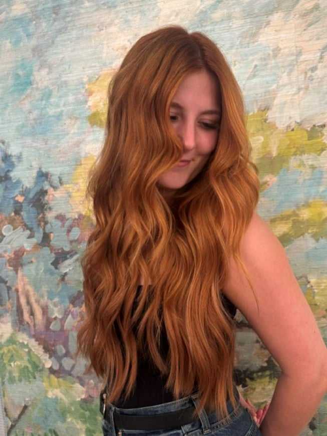 Young woman with long red hair and a black top stands before a colorful abstract background.