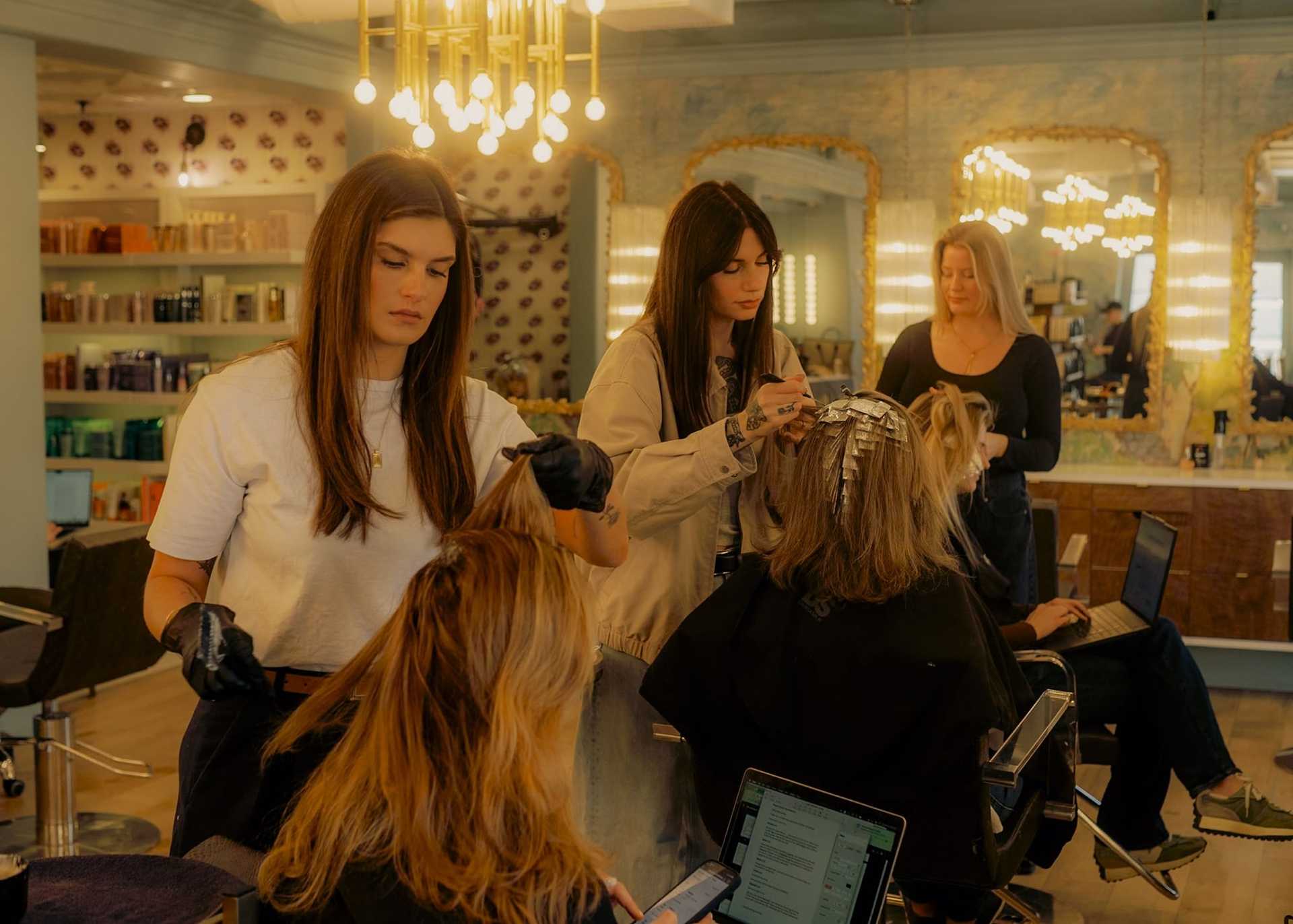 Hair salon with stylists coloring clients' hair; one client uses a laptop during the process.