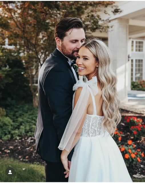Couple in wedding attire smiling outdoors, surrounded by greenery.